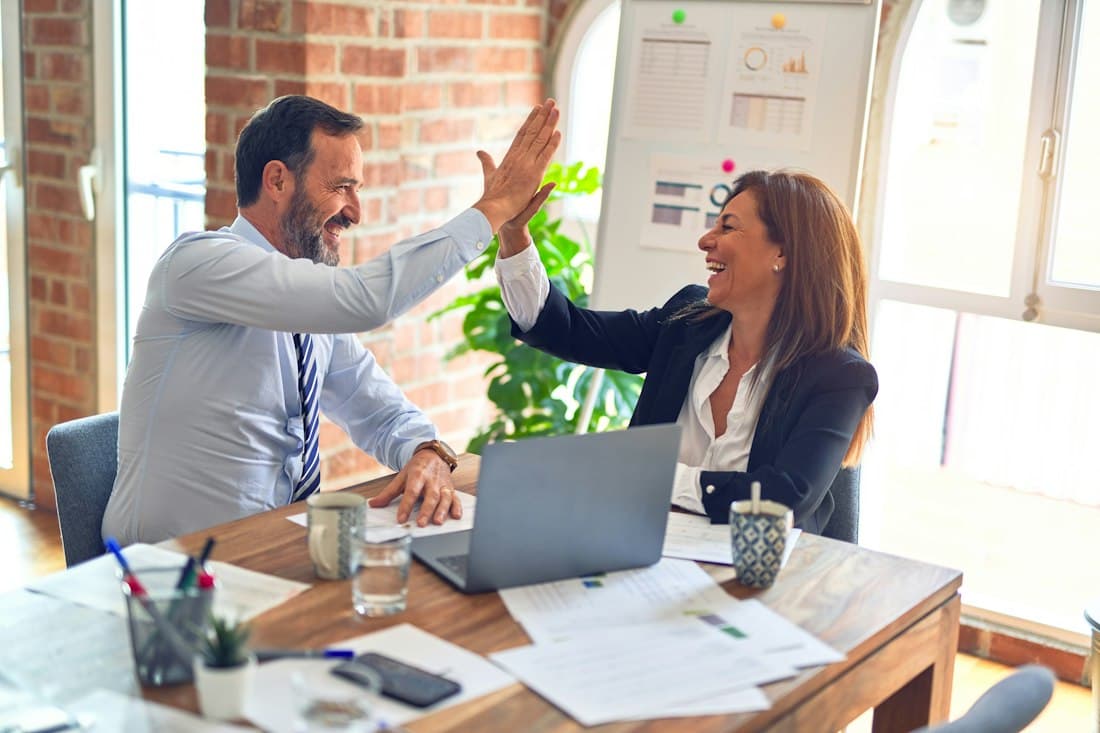 Two coworkers high-five across a desk with a laptop and paperwork in a bright office.
