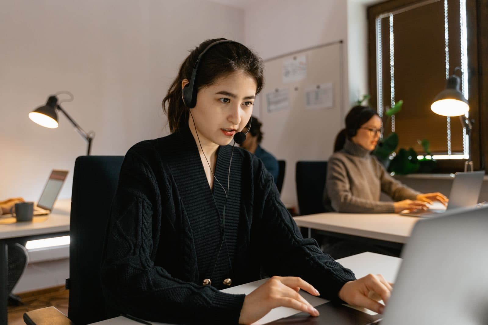 A focused assistant wearing a headset works on her laptop in a bright, lamp-lit workspace.
