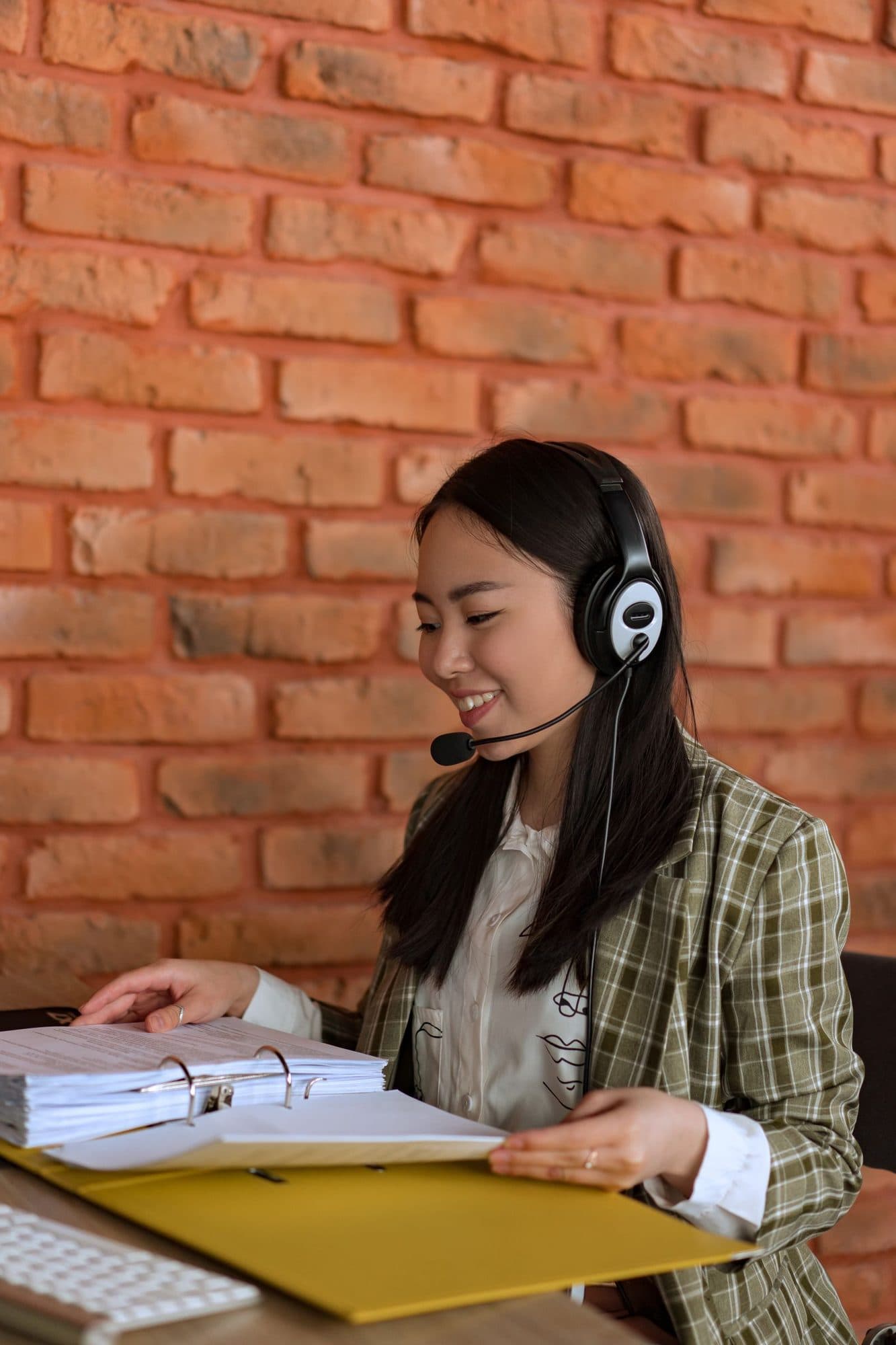 A smiling assistant wearing a headset reviews a binder of documents at a desk against a brick wall.