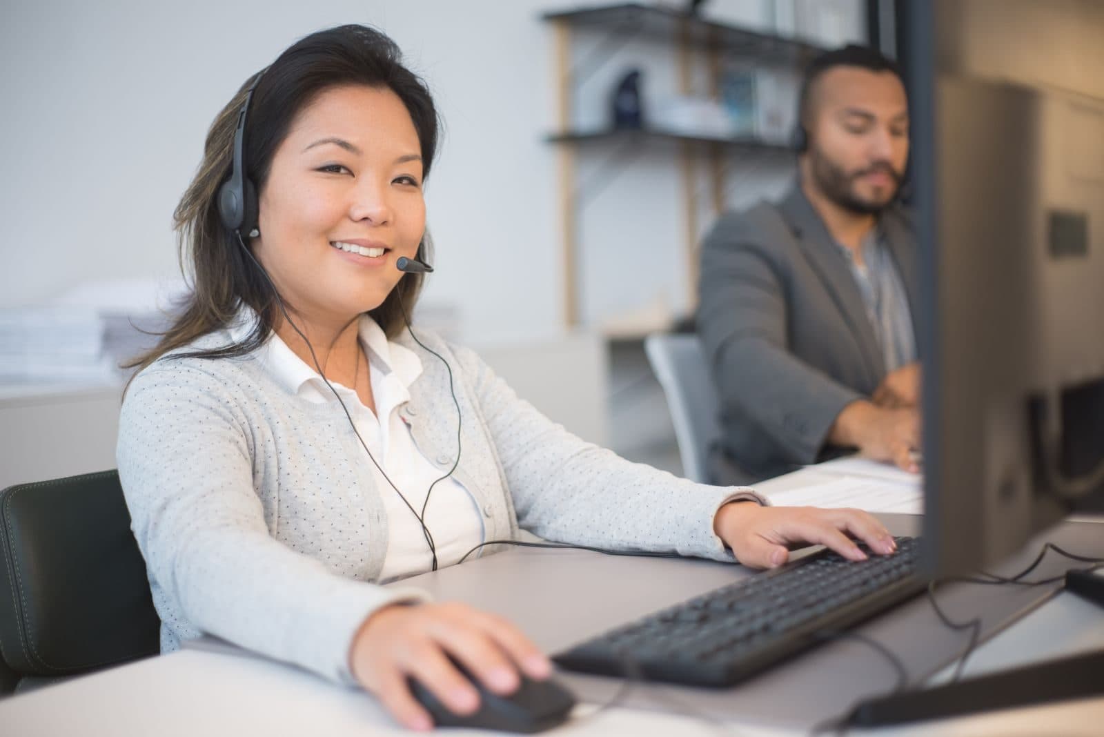 A smiling assistant wearing a headset works at her computer, with a colleague in the background.