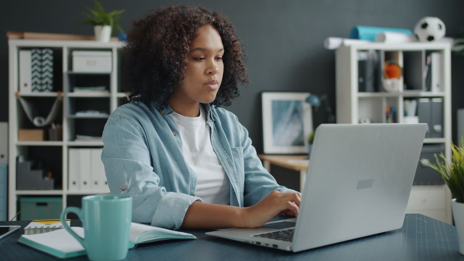 A woman with curly hair works on her laptop at a tidy home office desk with a mug and notebook.