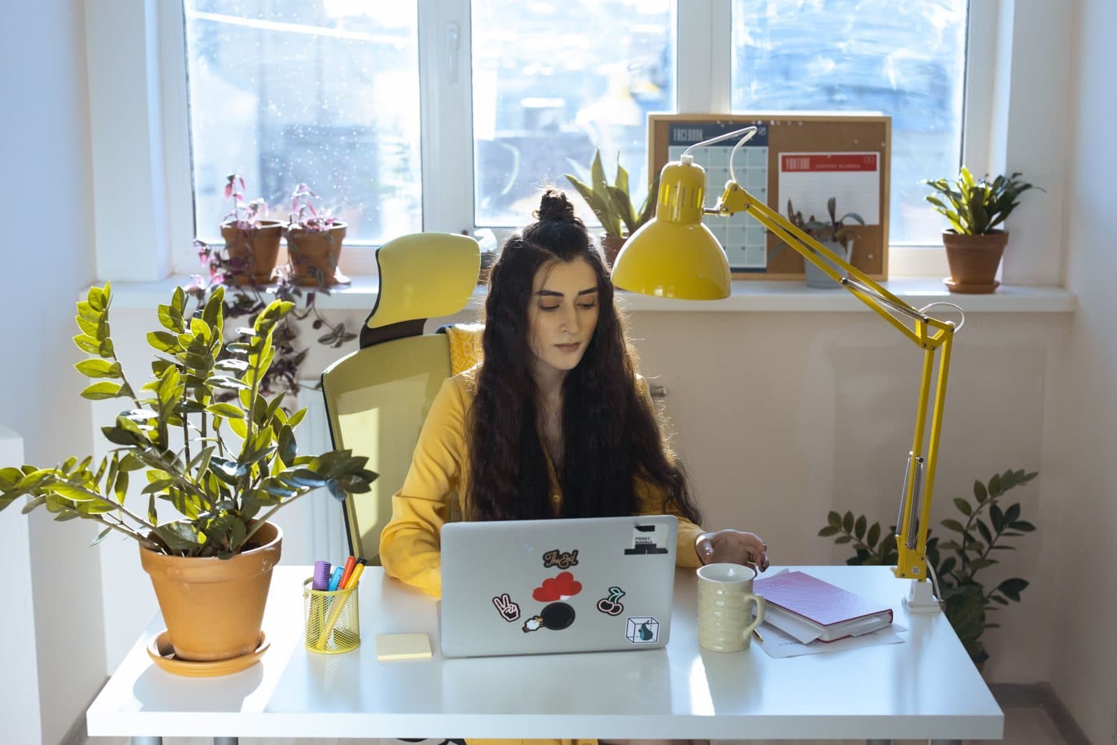 A woman in a yellow jacket works on her laptop at a bright desk surrounded by houseplants.