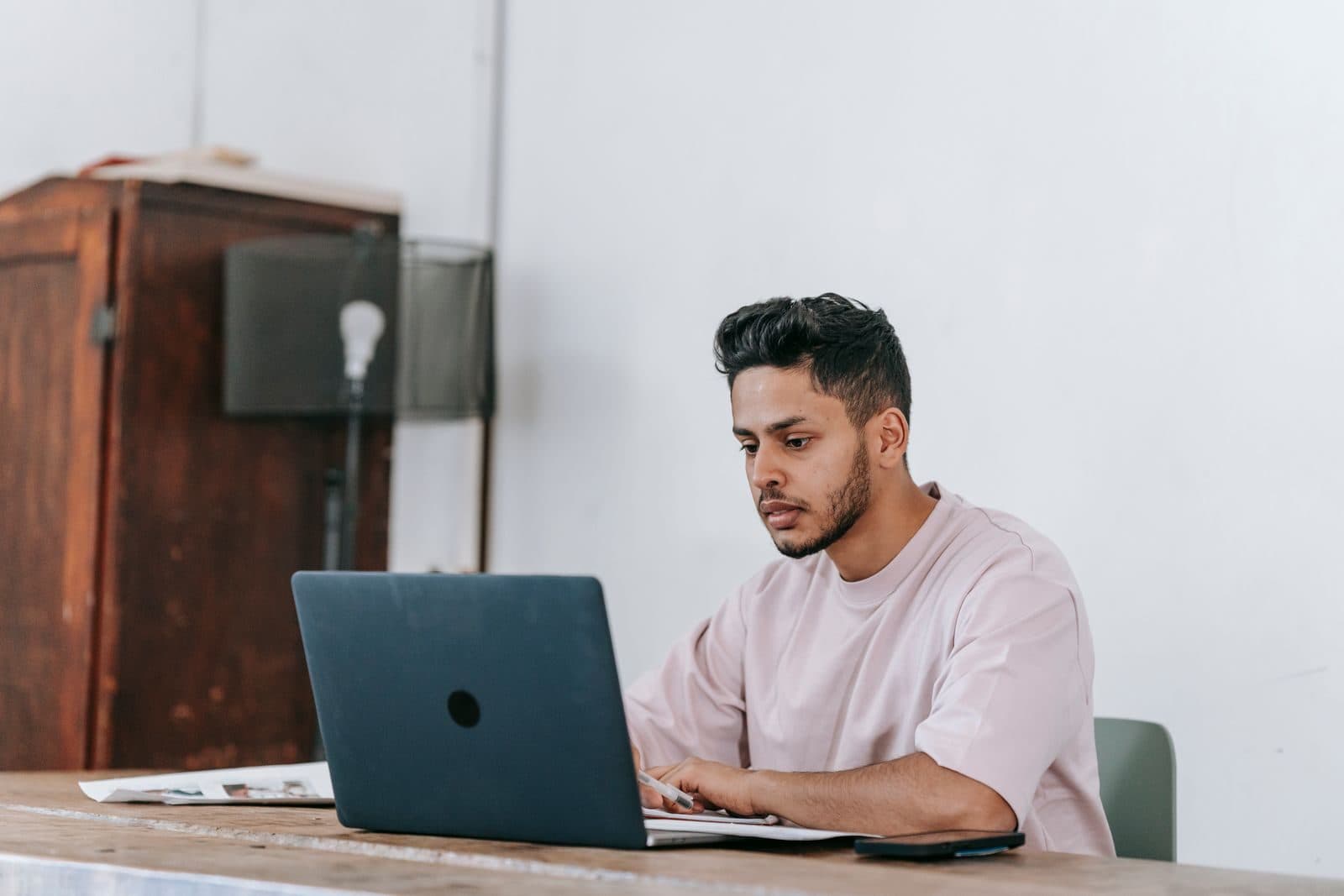 A young Latino professional concentrates on his laptop at a wooden table in a bright room.