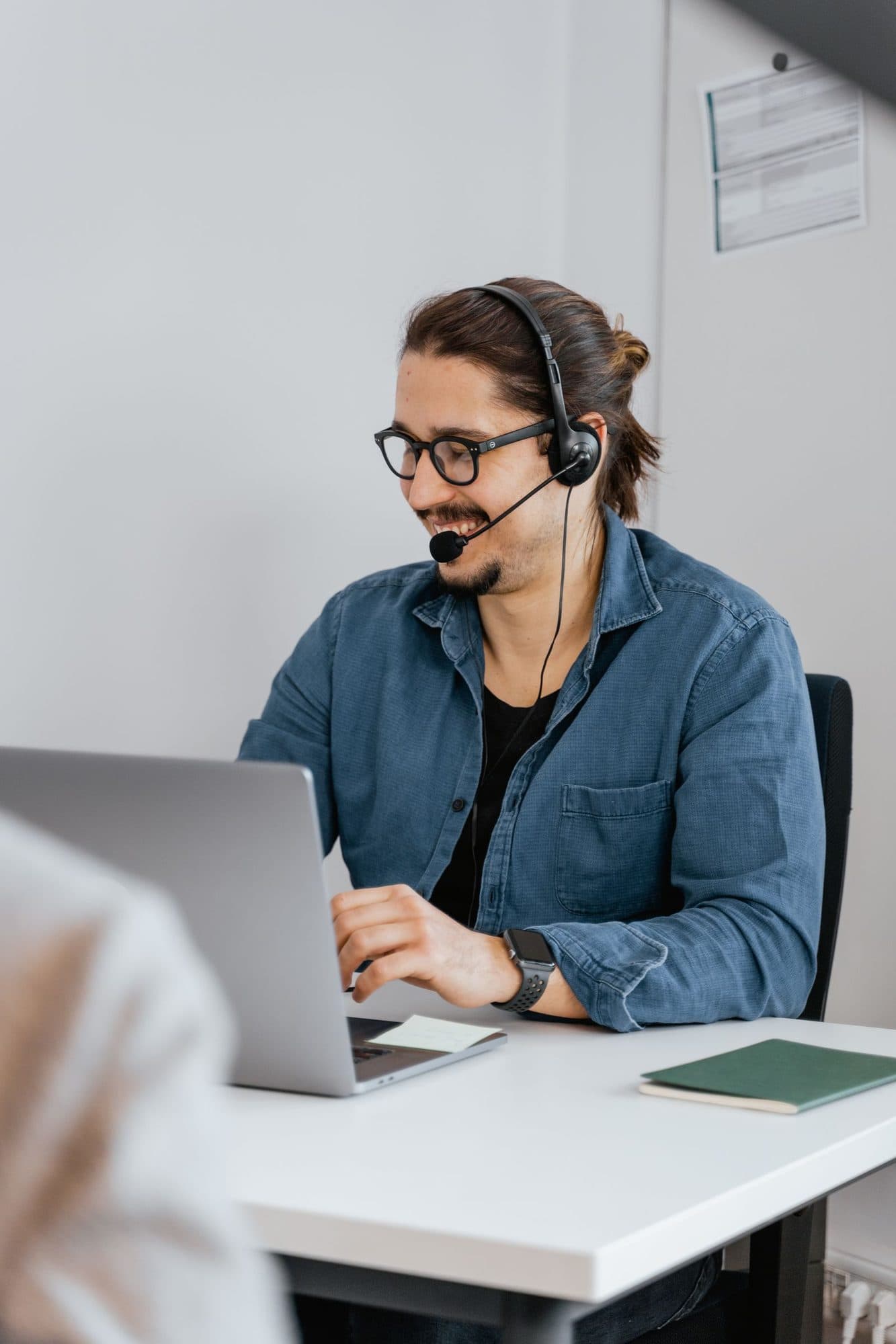 A smiling bearded professional in glasses and a denim shirt talks on a headset while working on his laptop.