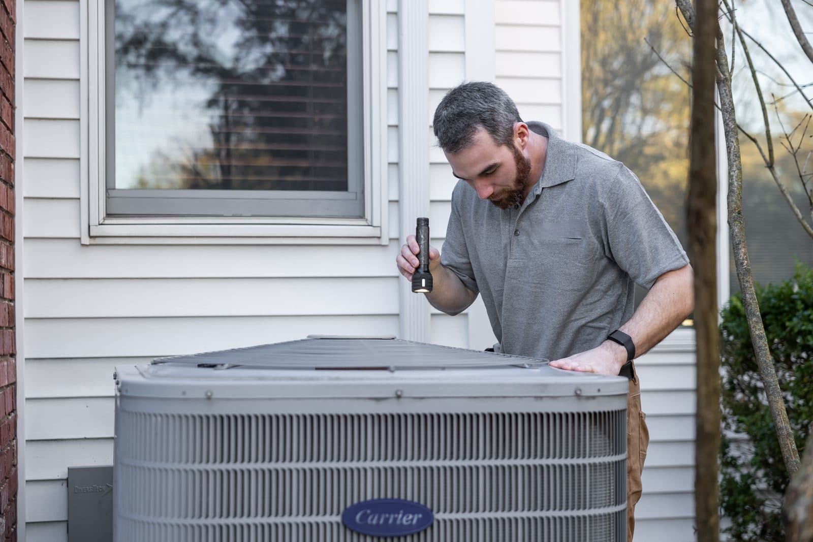 An HVAC technician uses a flashlight to inspect an outdoor air-conditioning unit beside a white-sided house.