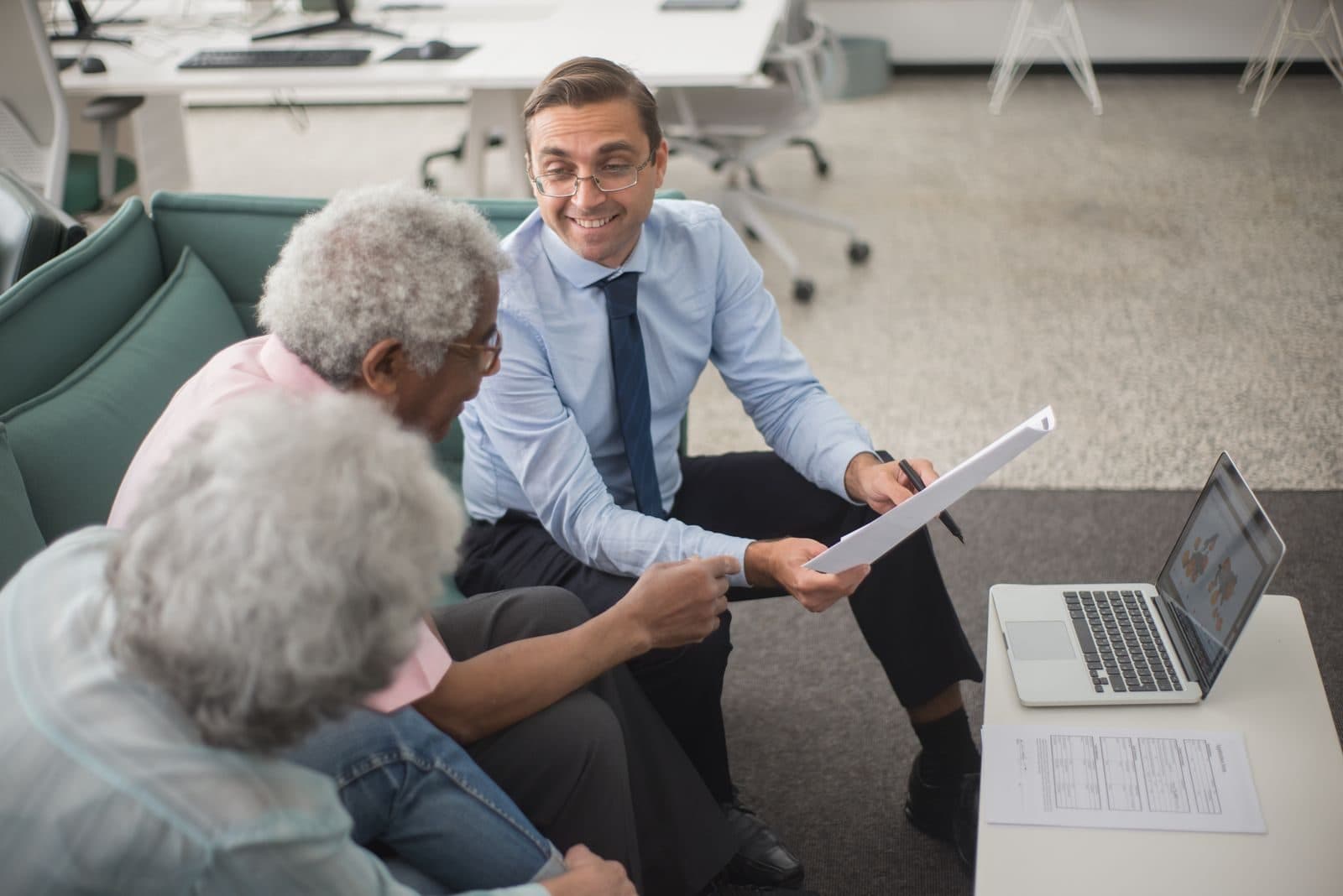 A smiling agent in a shirt and tie reviews paperwork with an older couple in a bright office.