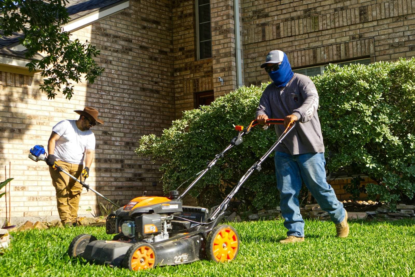 A two-person lawn crew mows and trims the yard in front of a brick house.