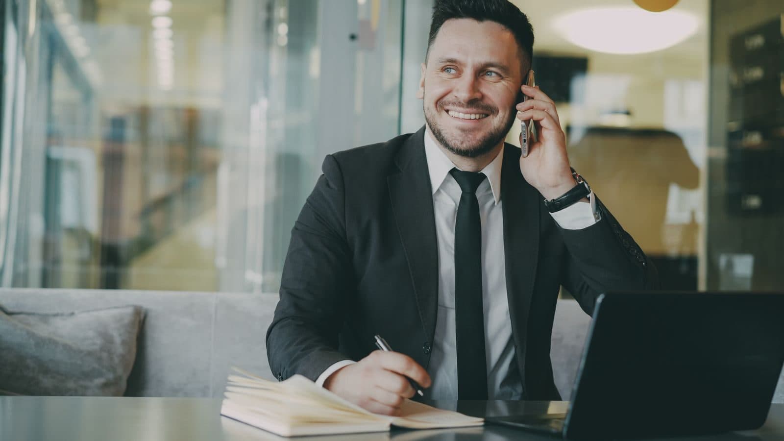 A smiling business owner in a suit takes a phone call while making notes at his desk.