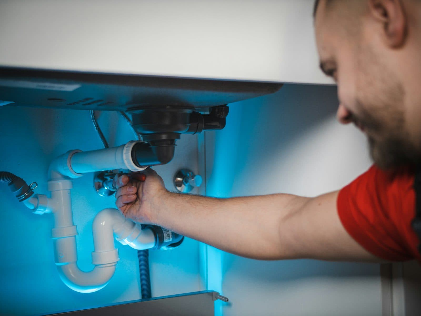 A smiling plumber reaches under a kitchen sink to tighten the drain pipes.