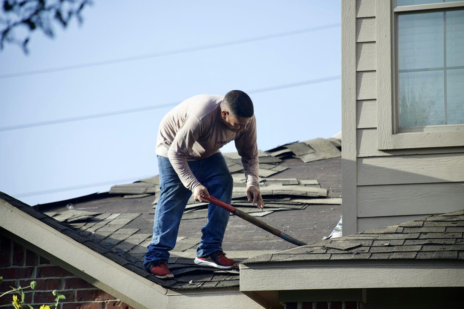 A roofer strips worn shingles from a residential roof on a clear day.