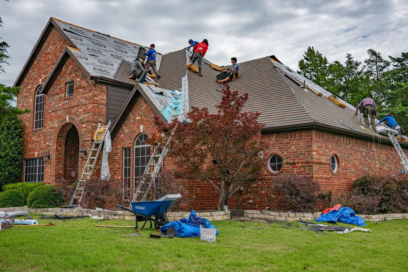 A roofing crew replaces shingles on a large brick house, with ladders and tarps on the lawn.