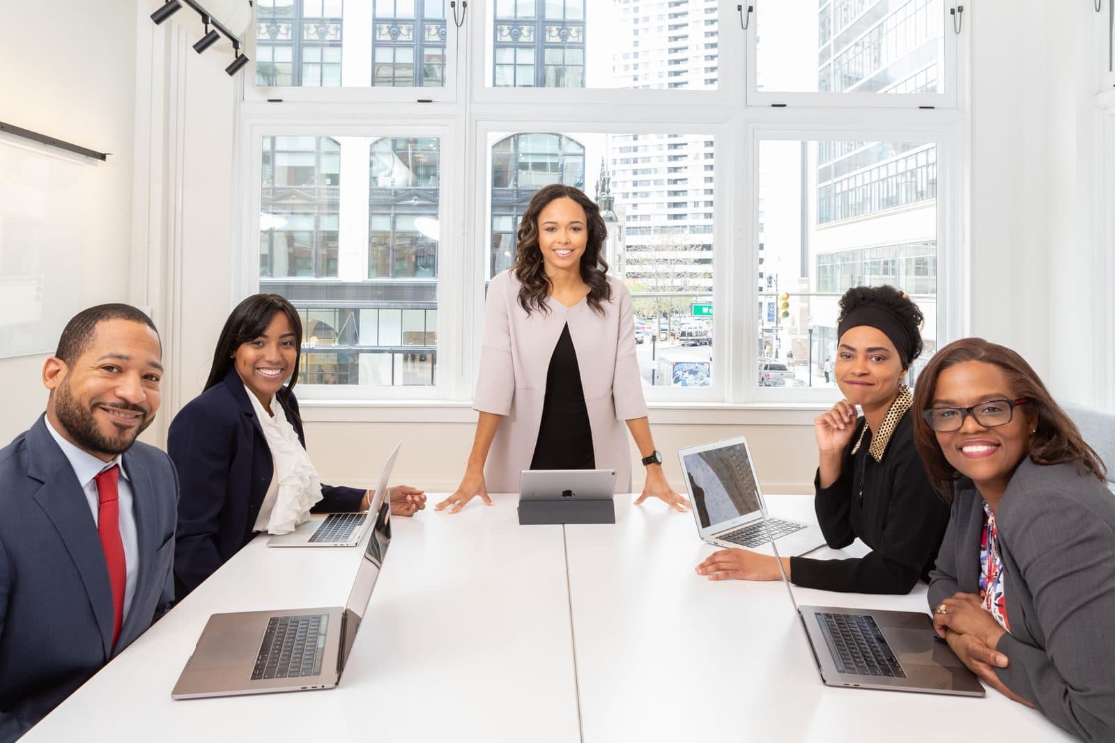 A professional team smiles around a conference table with laptops as a leader stands at the head.