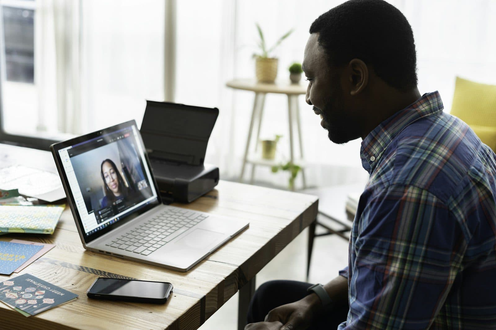 A smiling man in a plaid shirt talks with a woman on a laptop video call at his desk.