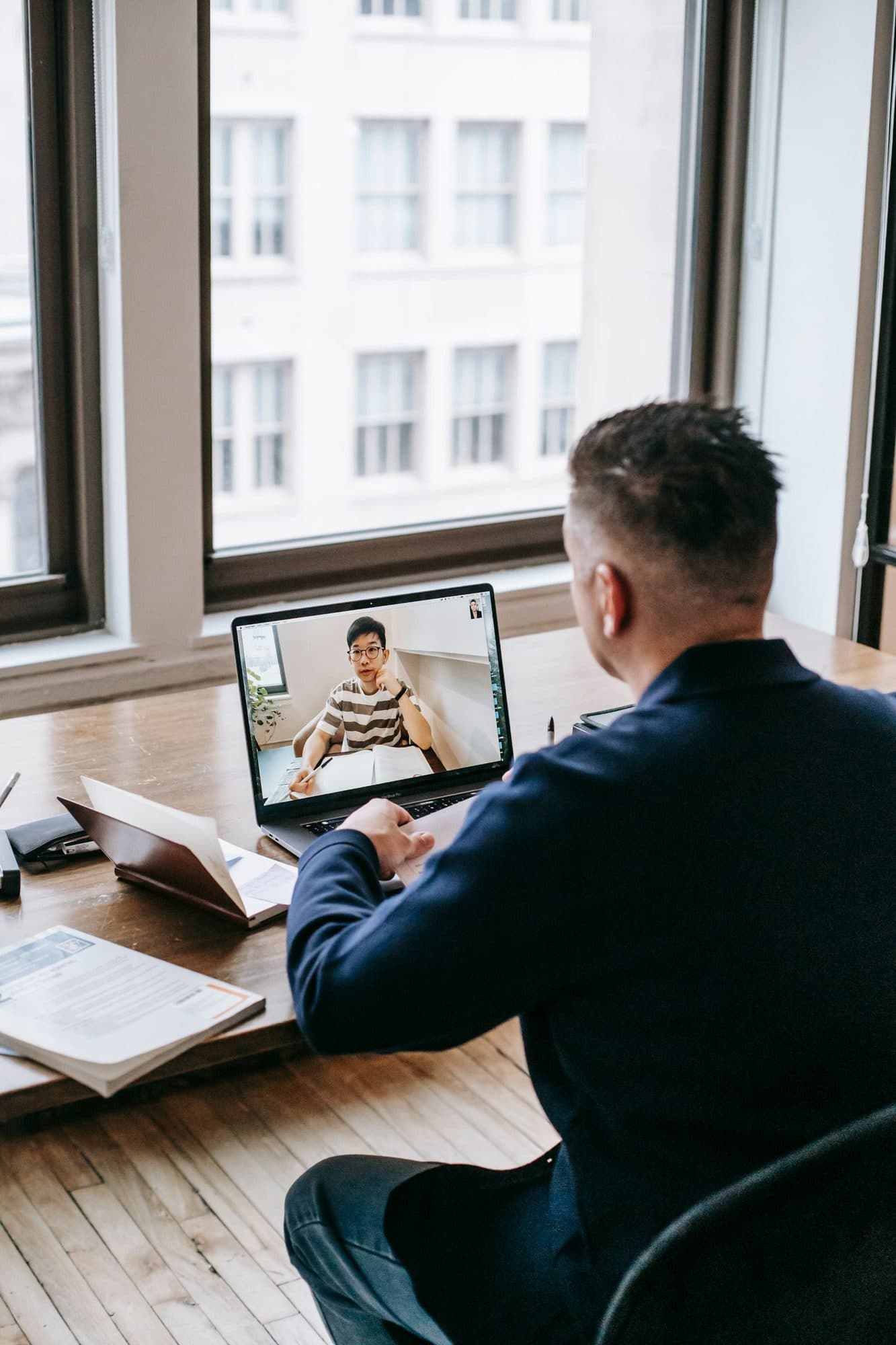 A manager in a navy sweater talks with a colleague on a laptop video call at a desk by a large window.