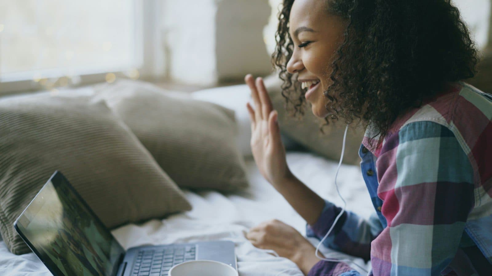 A smiling woman with curly hair waves hello to someone on a laptop video call.
