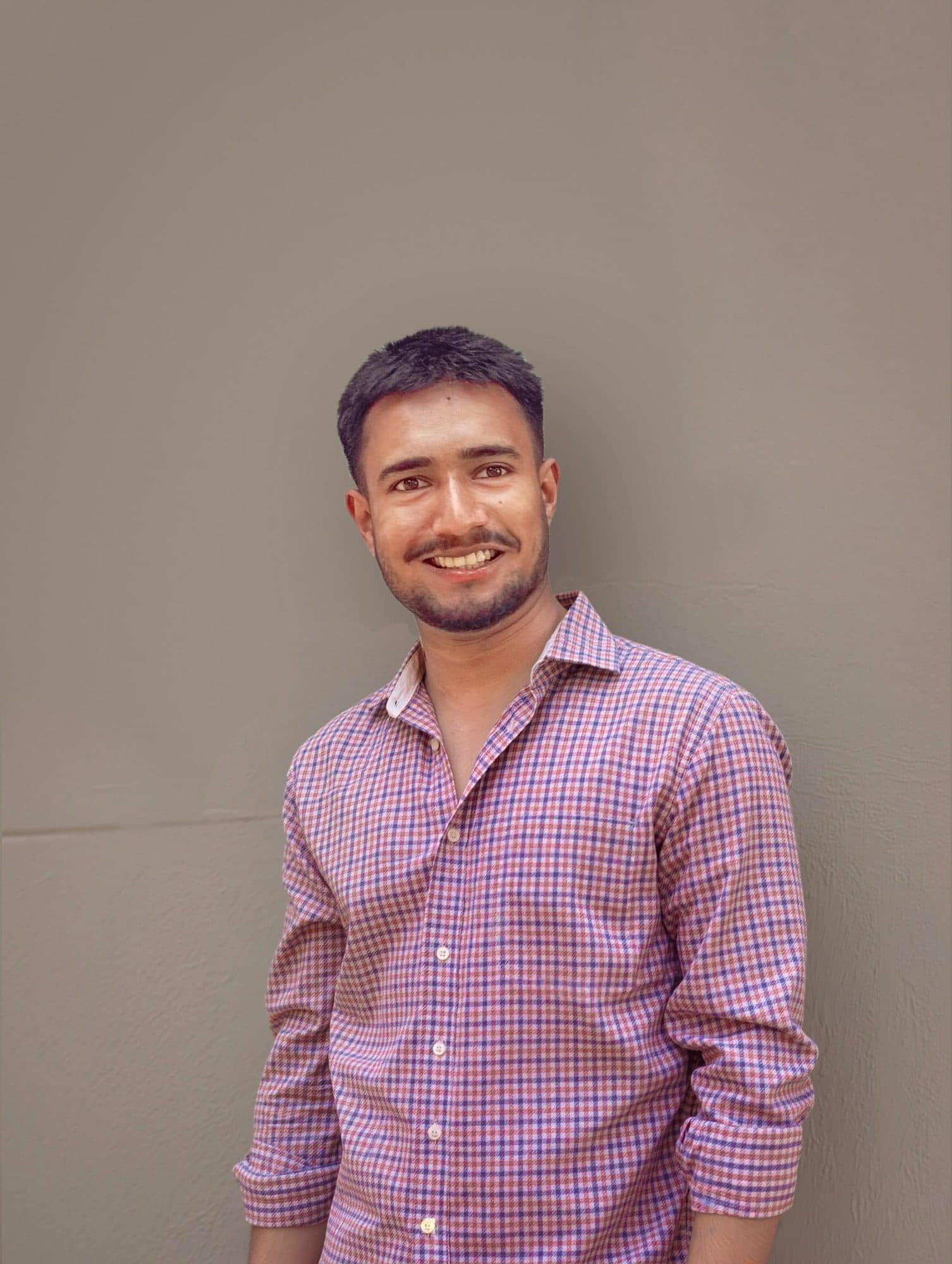 Portrait of a smiling young man in a checkered shirt against a neutral gray wall.