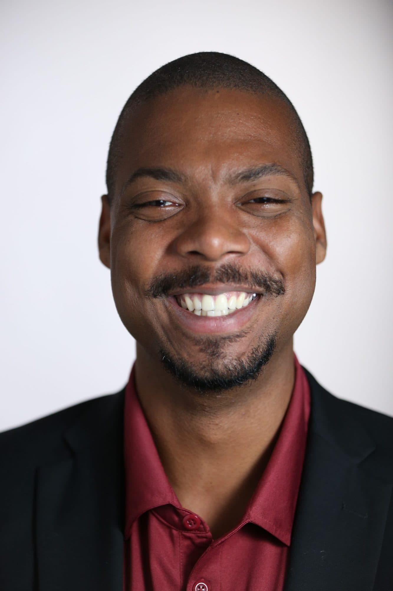 Portrait of a broadly smiling man in a dark blazer against a light background.