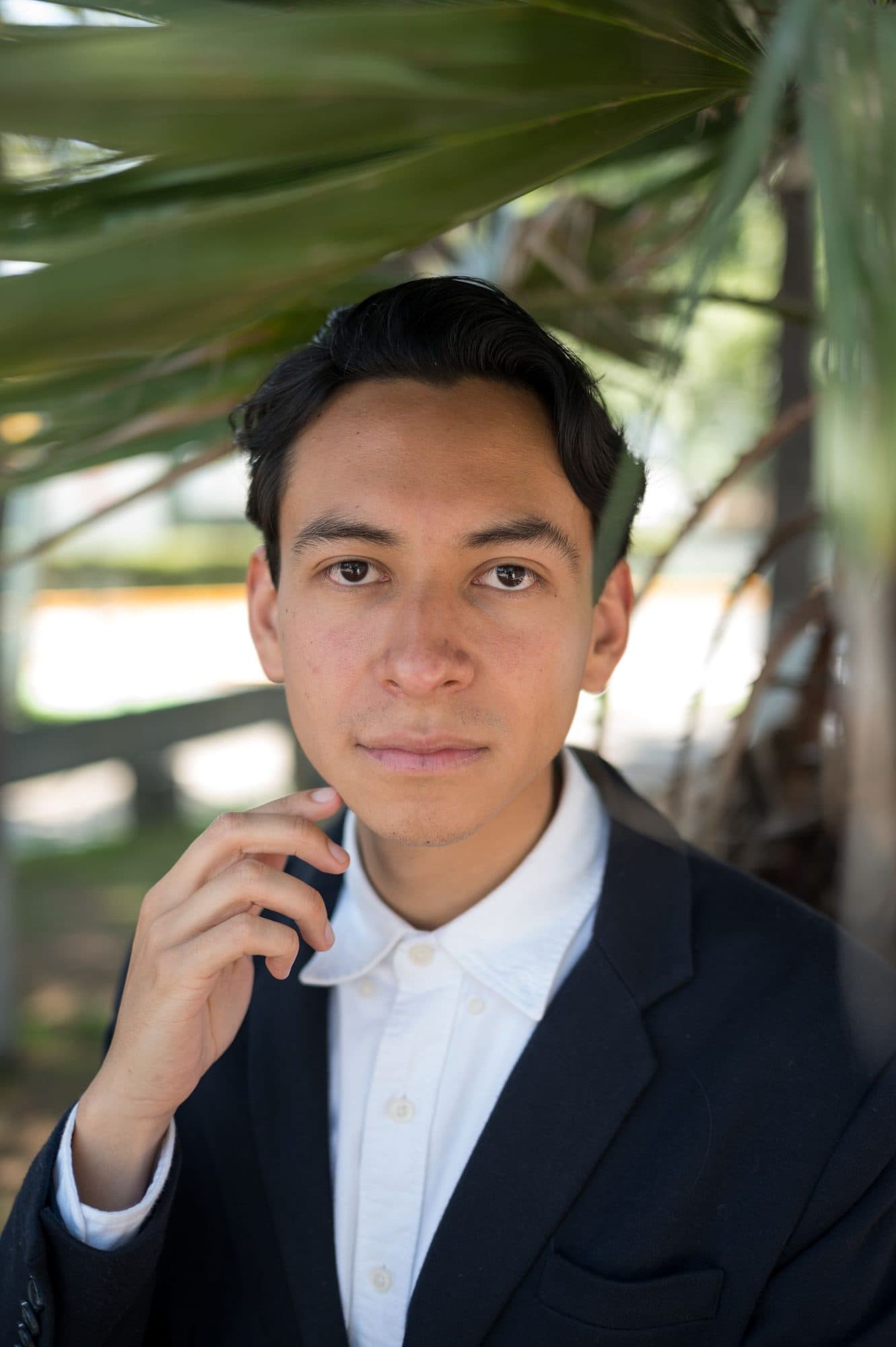 Portrait of a young man in a navy blazer and white shirt, framed by palm leaves.