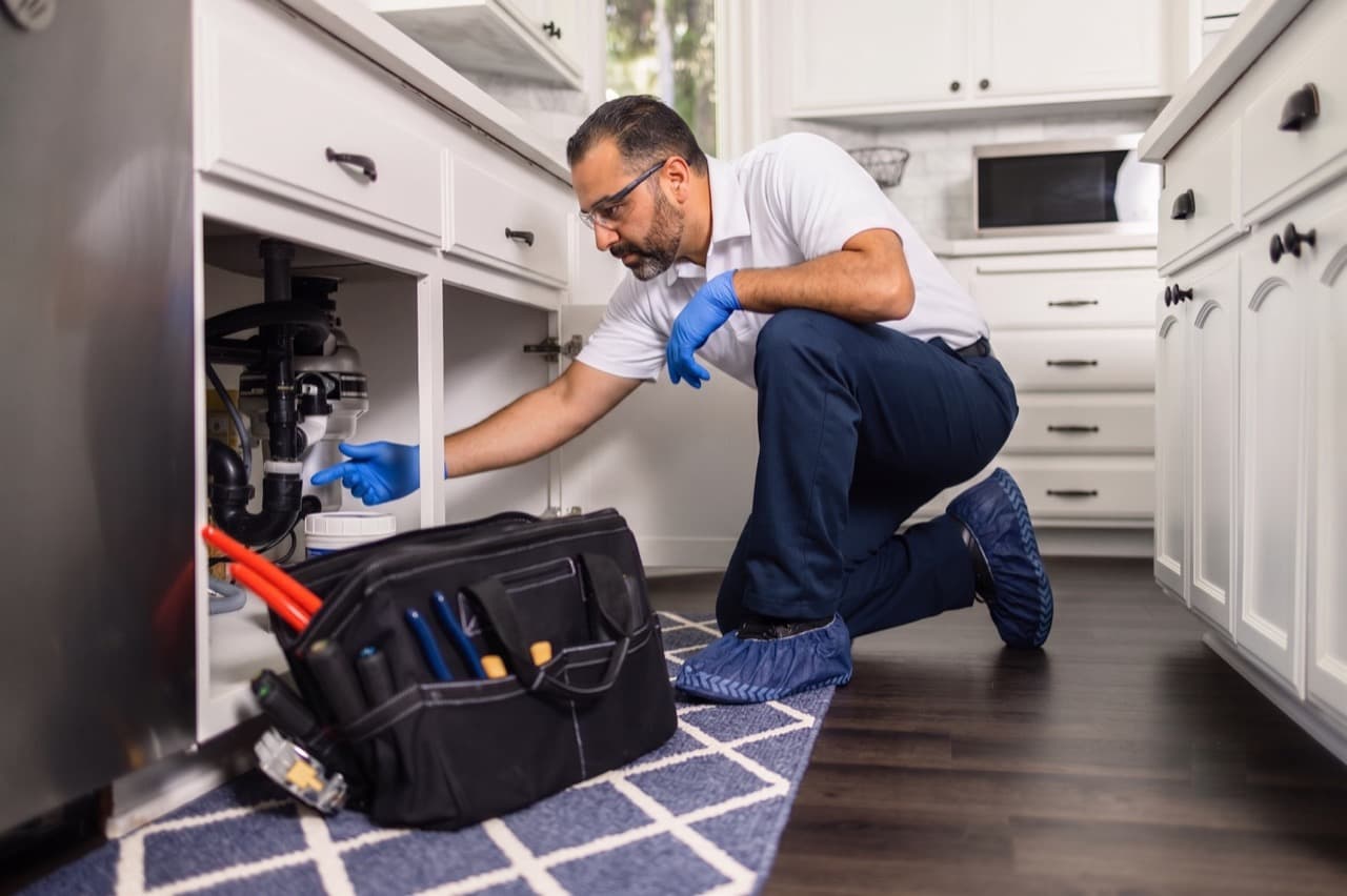 Plumber working under a kitchen sink with a tool bag.